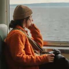 En ung kvinna med kamera sitter ensam i en buss och blickar ut över vattnet, en känsla av ensamhet i Sverige.