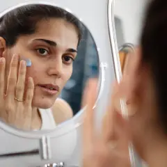 Kvinna applicerar en blå kräm i ansiktet framför spegeln, en del av hennes hudvårdsrutin för ansiktsbehandling mot acne.