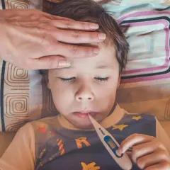 En hand känner på ett barns panna medan ett feberbarn har en termometer i munnen.