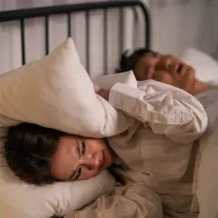 Kvinna med kudde över huvudet, plågad av ljud. En person sover bredvid. Känslighet för ljud kan vara en autism symptom.