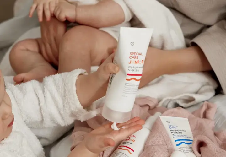 En bebis med torr hud får mjukgörande hudkräm på handen. Två tuber med "Special Care Junior" syns.