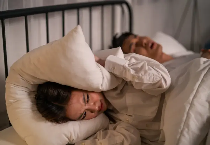 Kvinna med kudde över huvudet, plågad av ljud. En person sover bredvid. Känslighet för ljud kan vara en autism symptom.