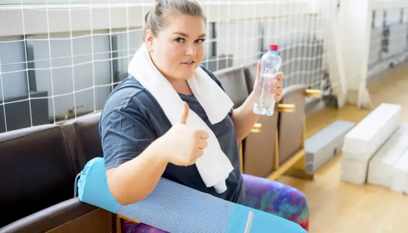 En kvinna med en handduk runt halsen ger tummen upp med en vattenflaska i handen. Hon sitter med en blå träningsmatta, redo för konservativ behandling.