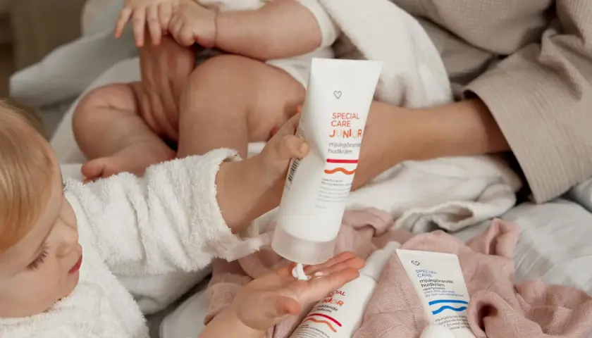 En bebis med torr hud får mjukgörande hudkräm på handen. Två tuber med "Special Care Junior" syns.