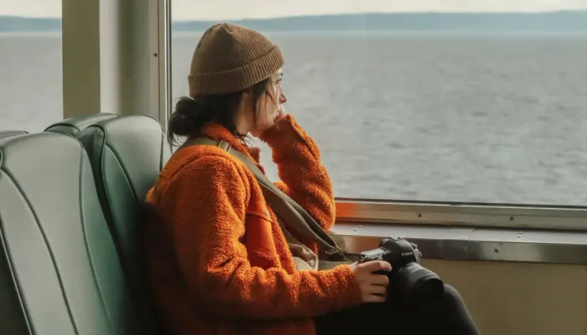En ung kvinna med kamera sitter ensam i en buss och blickar ut över vattnet, en känsla av ensamhet i Sverige.