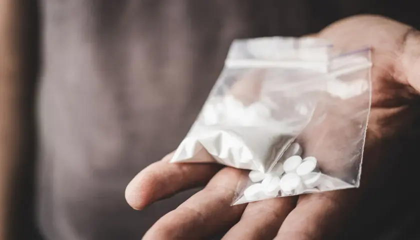 En hand håller en plastpåse med vitt pulver och tabletter, potentiellt crystal meth.