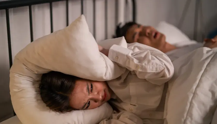 Kvinna med kudde över huvudet, plågad av ljud. En person sover bredvid. Känslighet för ljud kan vara en autism symptom.
