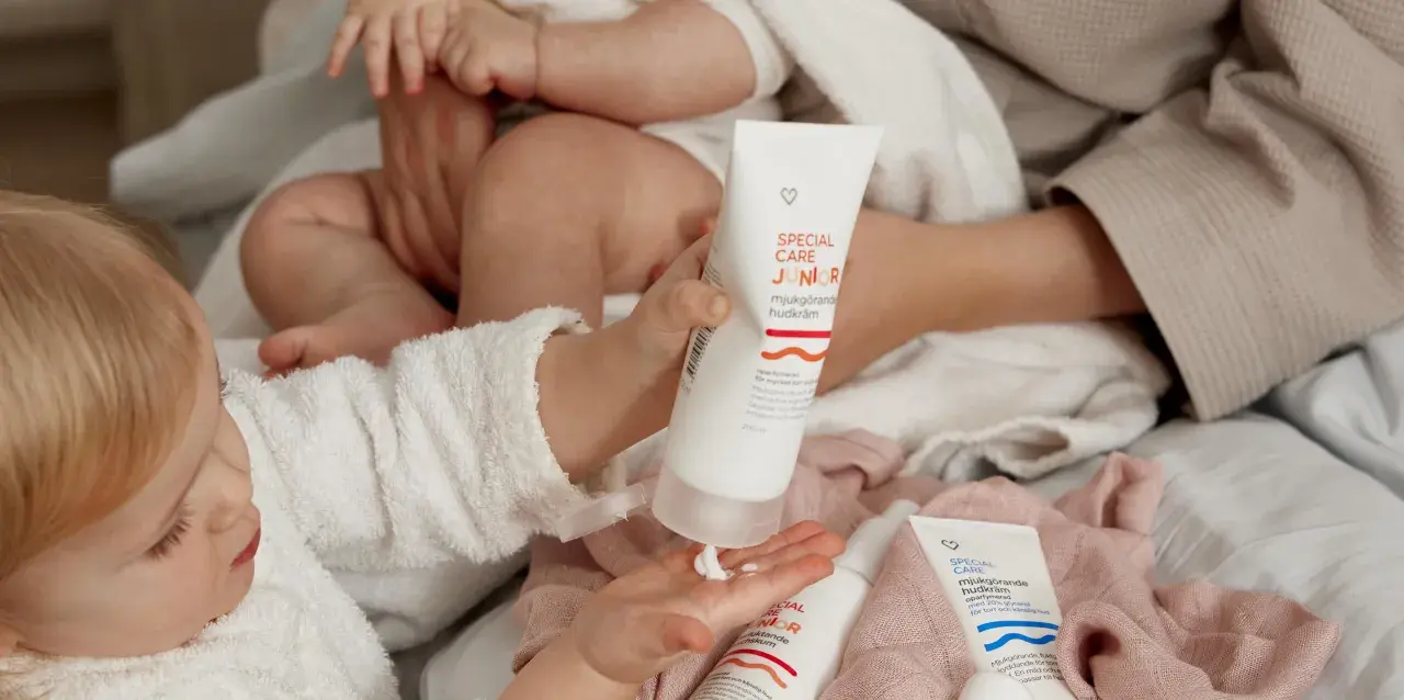 En bebis med torr hud får mjukgörande hudkräm på handen. Två tuber med "Special Care Junior" syns.