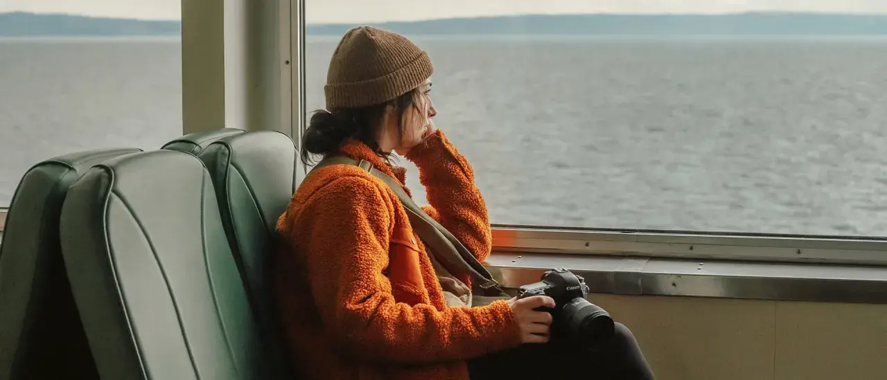 En ung kvinna med kamera sitter ensam i en buss och blickar ut över vattnet, en känsla av ensamhet i Sverige.