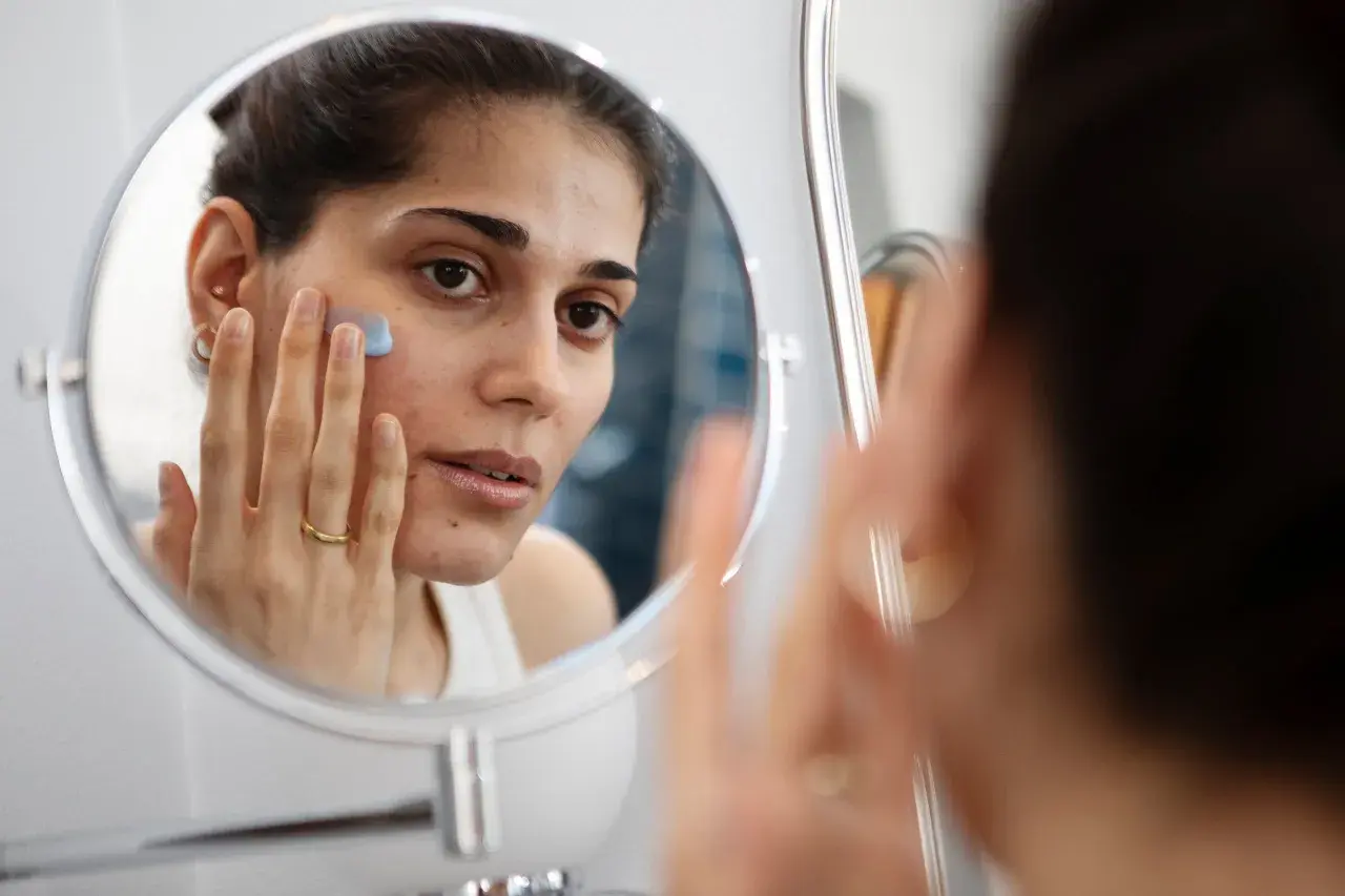Kvinna applicerar en blå kräm i ansiktet framför spegeln, en del av hennes hudvårdsrutin för ansiktsbehandling mot acne.