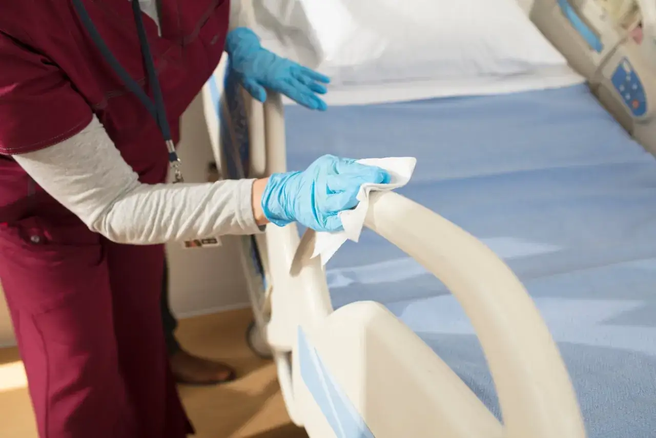A person in blue gloves wipes down a hospital bed rail with a white cloth.