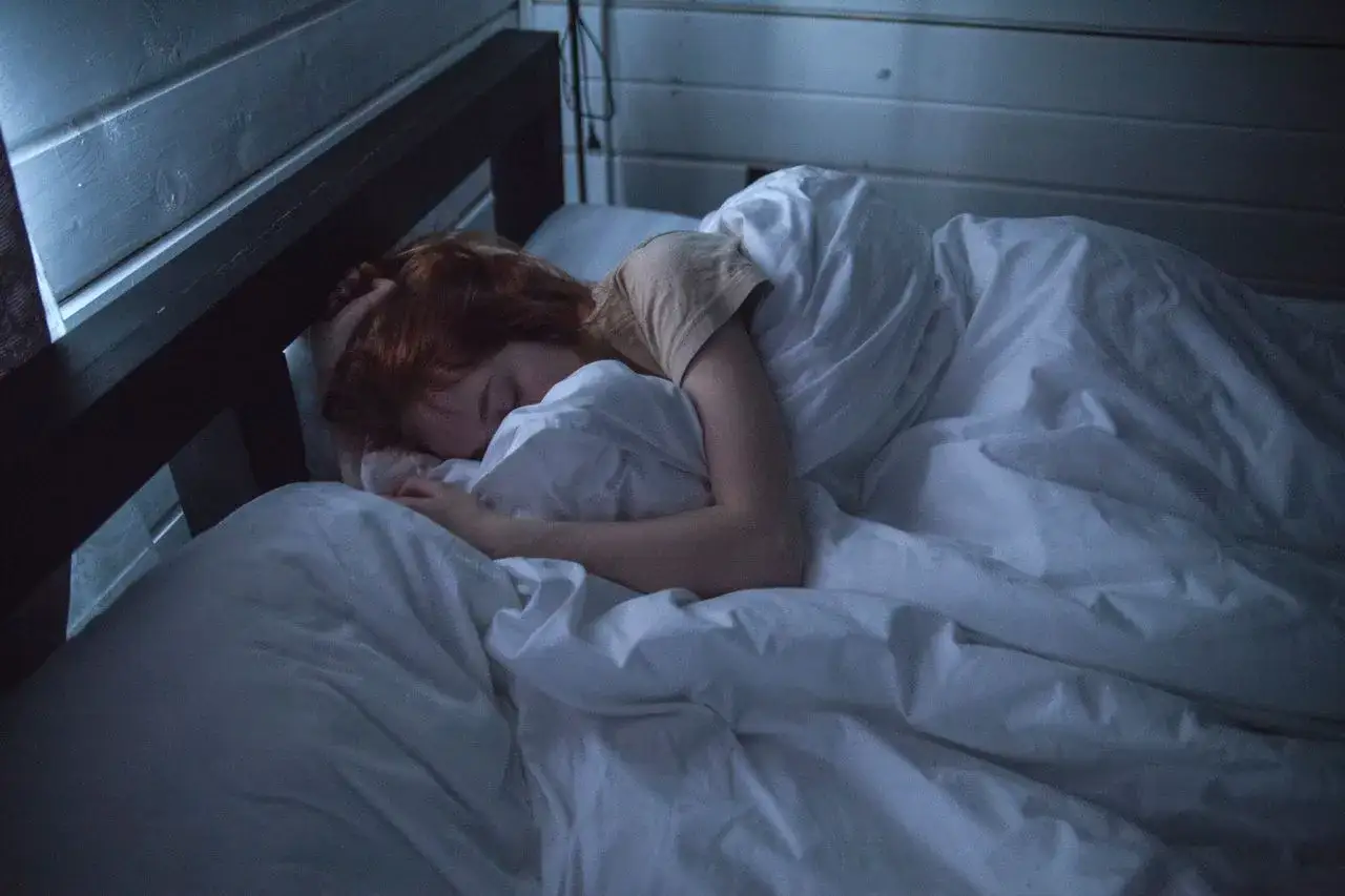 En person med r&ouml;tt h&aring;r sover ensam i en s&auml;ng. M&ouml;rkret och den vita s&auml;ngen f&ouml;rst&auml;rker k&auml;nslan av ofrivillig ensamhet.