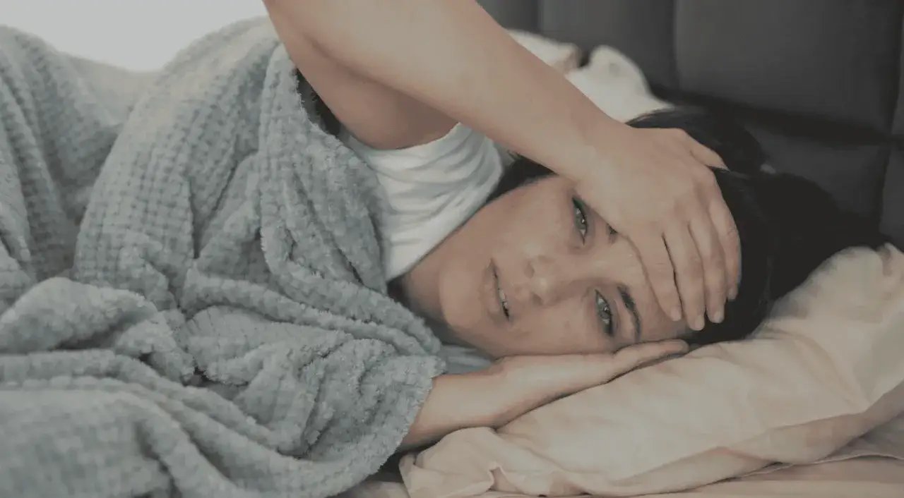 En kvinna ligger i s&auml;ngen, insvept i en filt, med handen mot pannan. Hon ser tr&ouml;tt och nedst&auml;md ut, vilket kan vara ett tecken p&aring; depression.