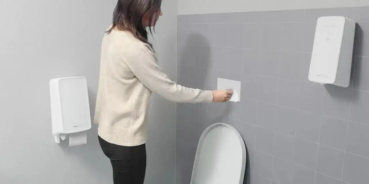 Kvinna torkar av spolknappen p&aring; toaletten f&ouml;r ytdesinfektion magsjuka. Pappershandduksh&aring;llare och ytdesinfektionsdispenser syns.